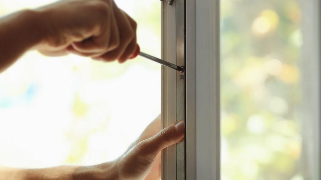 Close-up of a person's hand using a screwdriver to install or repair a window frame, with a blurred background suggesting a bright, sunny day outside, representing window installation and replacement.