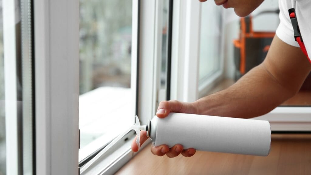 A person applying silicone caulk to the edge of a window frame to seal gaps, demonstrating a step in regular window maintenance for enhancing home insulation.