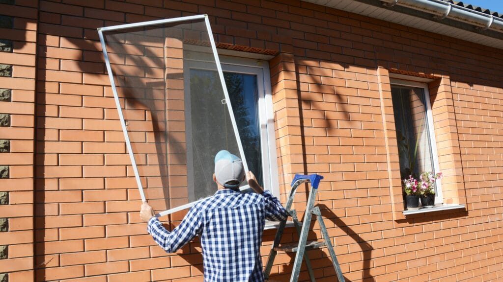 A person in a blue cap and plaid shirt installing a window screen on a sunny day, with the red brick exterior of a house providing a warm background.